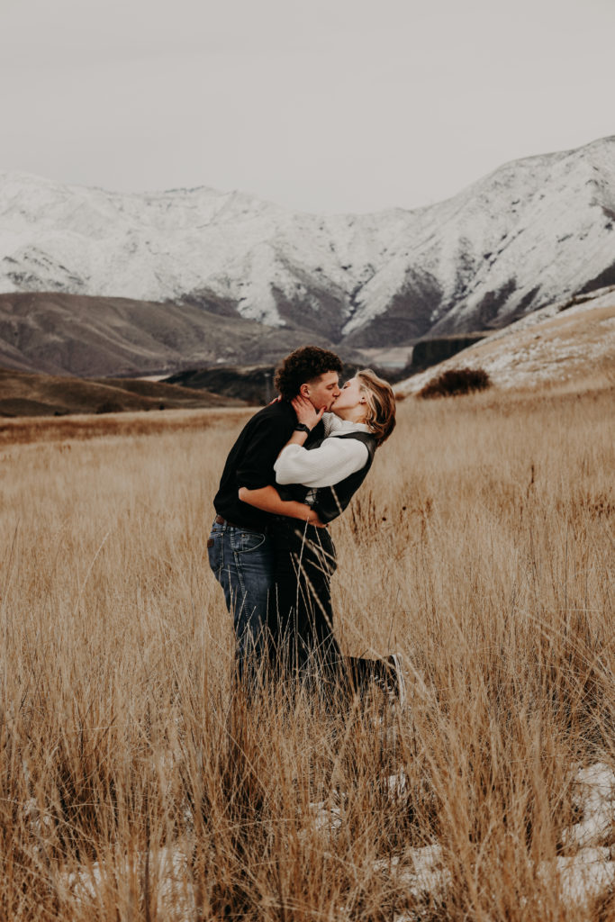 Intamate couples photography with guy leaning over girl and kissing in feild with mountain backdrop. 