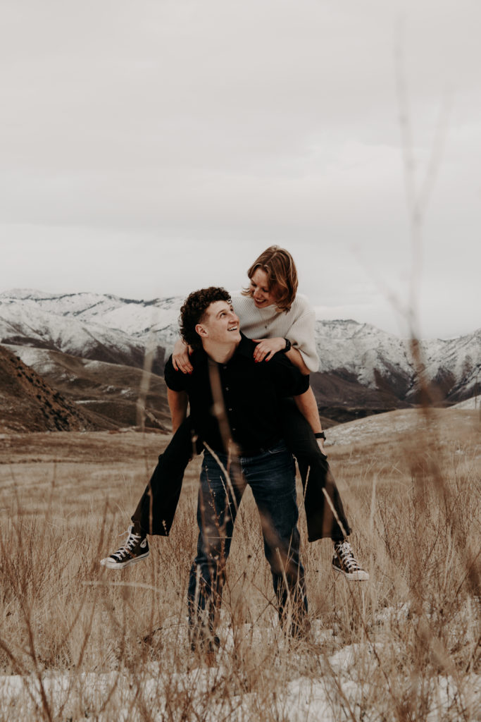 Couples posing piggy back ride in feild with mountain background. 